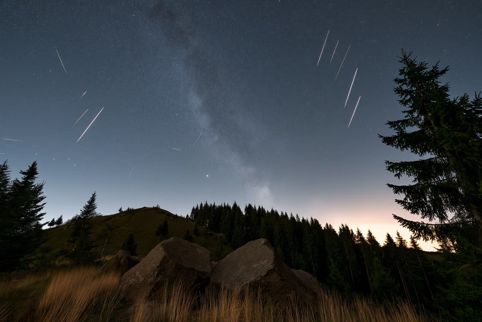 Noćno nebo nad tamnim njemačkim krajolikom s meteorskim prugama i slabim pojasom zvijezda iznad glave.